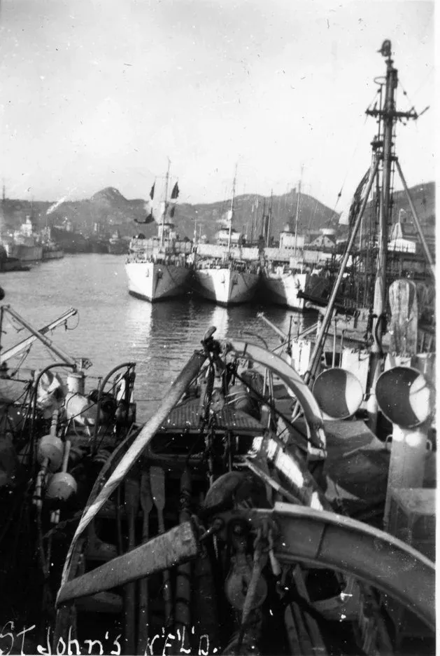 Two minesweepers and corvettes secured in harbour with destroyers, frigates, and a fleet of merchant ships in the background — photo taken from HMCS Wasaga