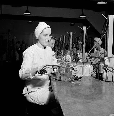 Women factory workers solder fuse boxes at a table in a munitions factory