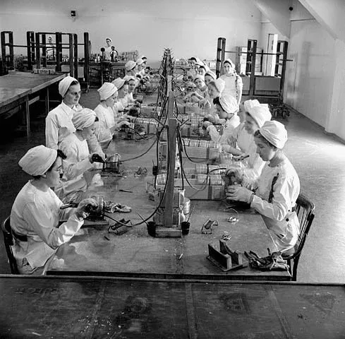 Women munitions workers solder fuse boxes at a long table in a munitions factory